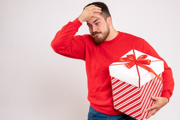 Vorderansicht des jungen mannes im roten hemd, das weihnachtsgeschenk im kasten auf weißer wand hält