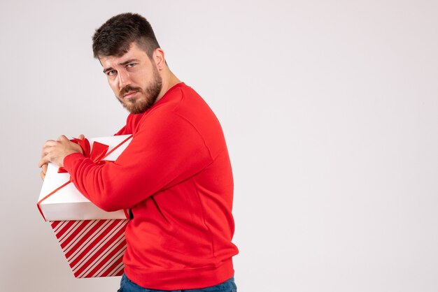 Vorderansicht des jungen Mannes im roten Hemd, das Weihnachtsgeschenk im Kasten auf weißer Wand hält