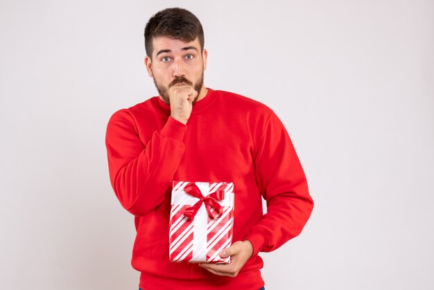 Vorderansicht des jungen Mannes im roten Hemd, das Weihnachtsgeschenk nervös auf weißer Wand hält