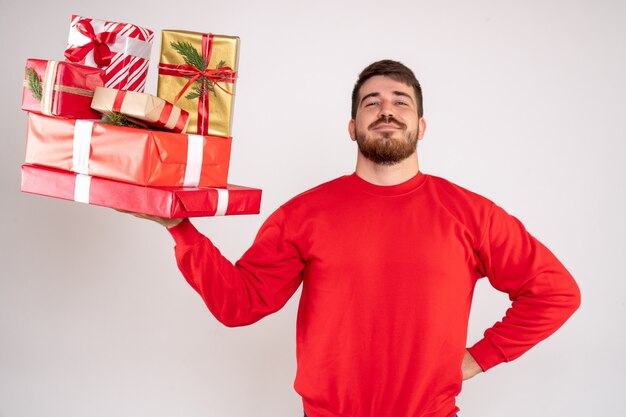 Vorderansicht des jungen Mannes im roten Hemd, das Weihnachtsgeschenke auf weißer Wand hält