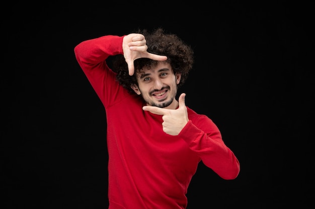 Vorderansicht des jungen Mannes im roten Pullover, der auf schwarzer Wand posiert und lächelt