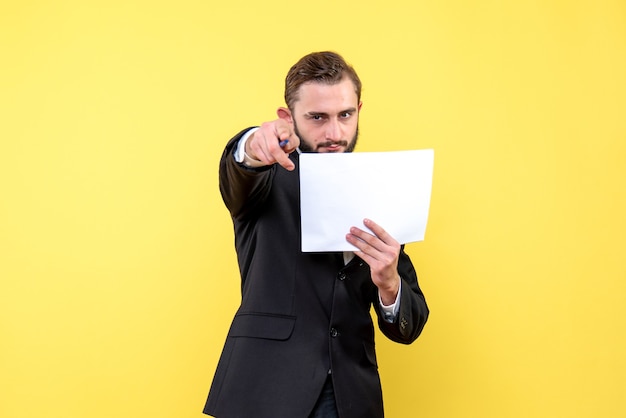 Vorderansicht des jungen Mannes im schwarzen Anzug, der mit einem Stift schaut und streng zeigt, der ein leeres Dokument auf Gelb hält