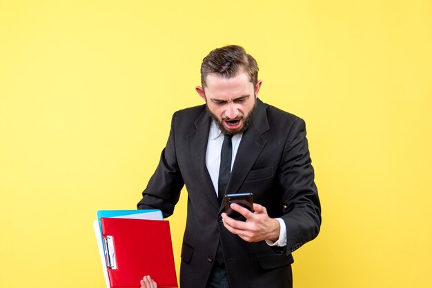 Vorderansicht des jungen Mannes im schwarzen Anzug leiden Blick auf ein Mobiltelefon und hält Ordner auf gelb