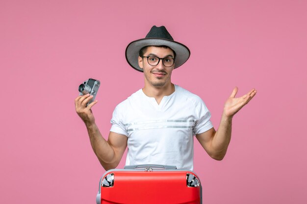 Vorderansicht des jungen Mannes im Urlaub mit roter Tasche mit Kamera an rosa Wand