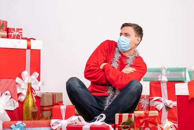 Vorderansicht des jungen Mannes in Maske, der um Weihnachtsgeschenke auf weißer Wand sitzt