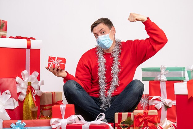 Vorderansicht des jungen Mannes in Maske, der um Weihnachtsgeschenke auf weißer Wand sitzt