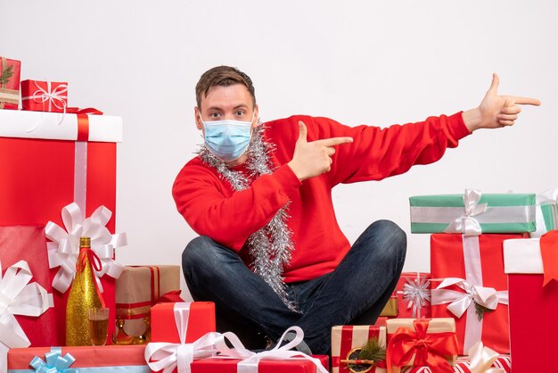 Vorderansicht des jungen Mannes in Maske, der um Weihnachtsgeschenke auf weißer Wand sitzt