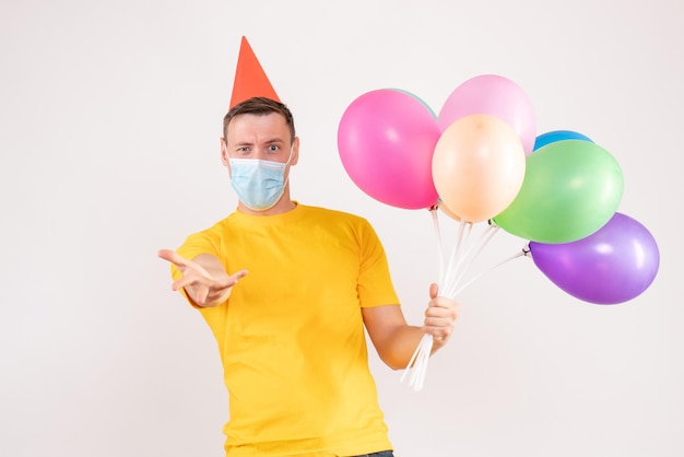 Vorderansicht des jungen Mannes mit bunten Luftballons in Maske auf weißer Wand