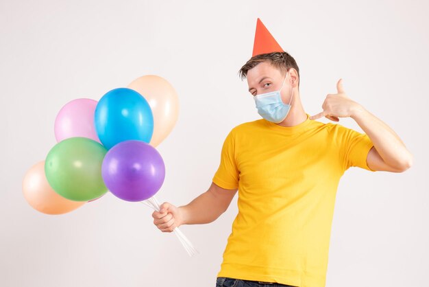 Vorderansicht des jungen Mannes mit bunten Luftballons in Maske auf weißer Wand