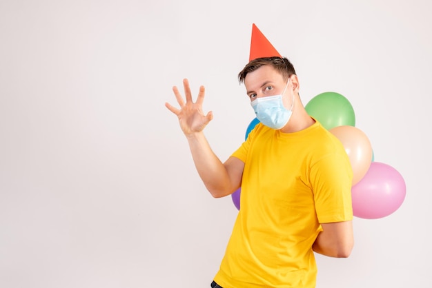 Vorderansicht des jungen Mannes mit bunten Luftballons in Maske auf weißer Wand