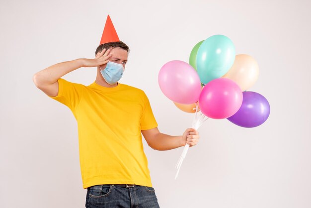 Vorderansicht des jungen Mannes mit bunten Luftballons in Maske auf weißer Wand