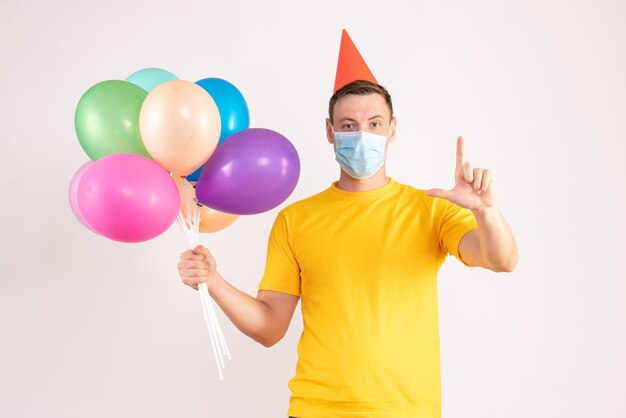 Vorderansicht des jungen Mannes mit bunten Luftballons in steriler Maske auf weißer Wand