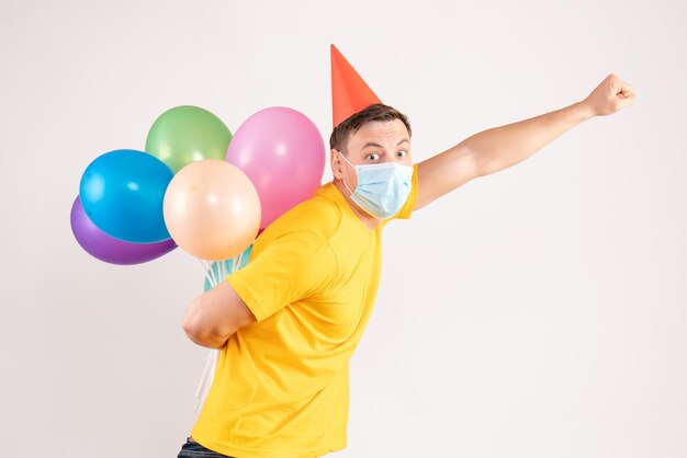 Vorderansicht des jungen Mannes mit bunten Luftballons in steriler Maske auf weißer Wand
