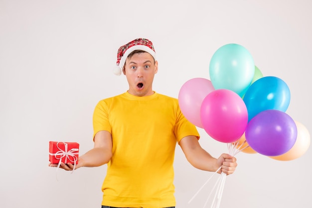 Vorderansicht des jungen Mannes mit bunten Luftballons und kleinem Geschenk auf weißer Wand