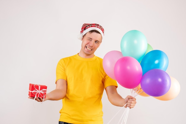 Kostenloses Foto vorderansicht des jungen mannes mit bunten luftballons und kleinem geschenk auf weißer wand