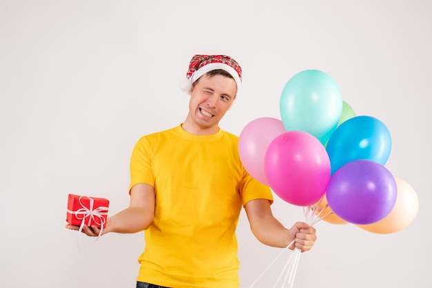 Vorderansicht des jungen Mannes mit bunten Luftballons und kleinem Geschenk auf weißer Wand