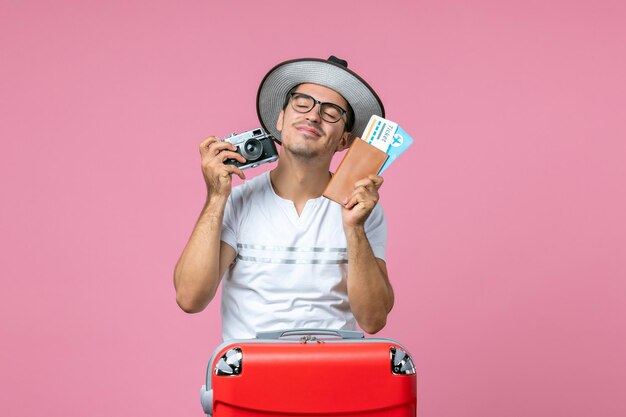 Vorderansicht des jungen Mannes mit Kamera und Flugtickets an der rosa Wand