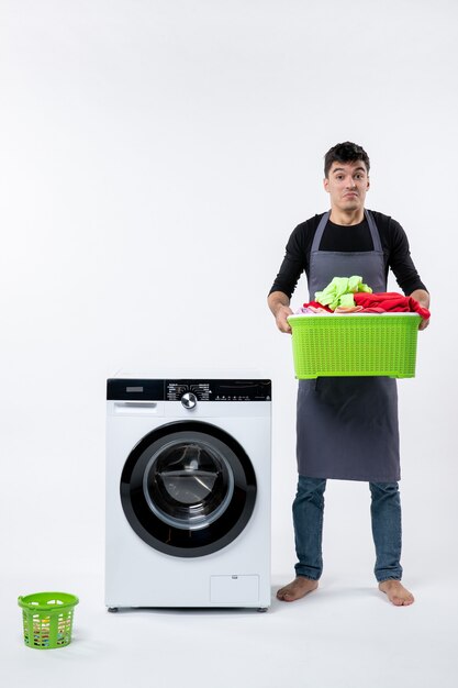 Vorderansicht des jungen Mannes mit schmutziger Kleidung und Waschmaschine auf weißer Wand