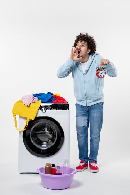 Vorderansicht des jungen Mannes mit Waschmaschine, die bis zum Ende des Waschens der Kleidung an der weißen Wand wartet