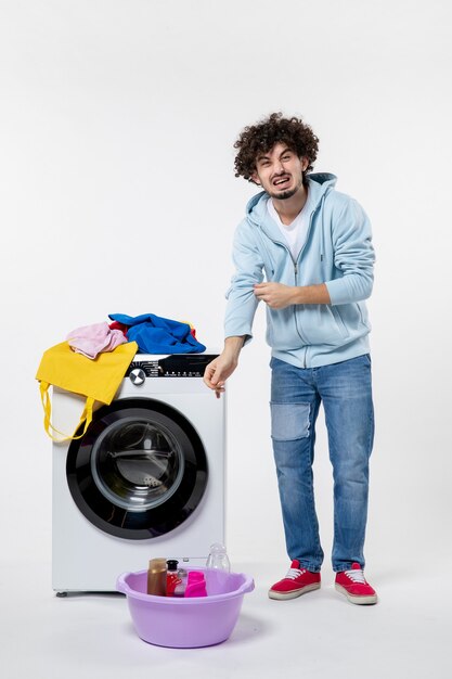 Vorderansicht des jungen Mannes mit Waschmaschine und schmutziger Kleidung auf weißer Wand