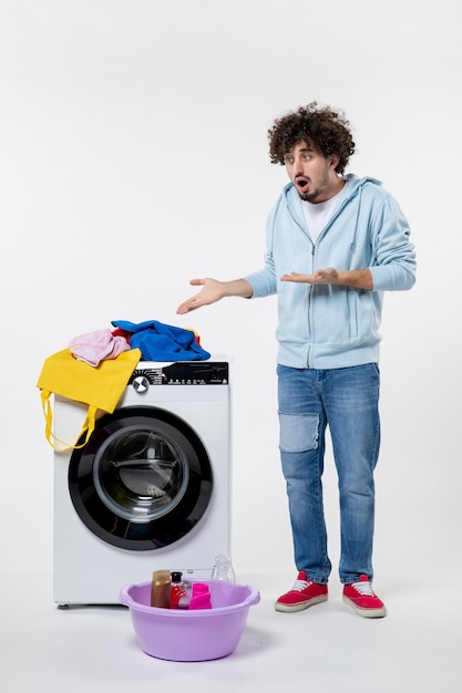 Vorderansicht des jungen Mannes mit Waschmaschine und schmutziger Kleidung auf weißer Wand