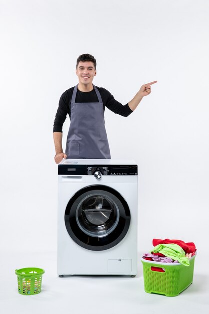 Vorderansicht des jungen Mannes mit Waschmaschine und schmutziger Kleidung auf weißer Wand