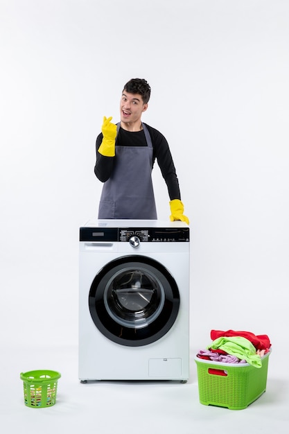 Vorderansicht des jungen Mannes mit Waschmaschine und schmutziger Kleidung auf weißer Wand