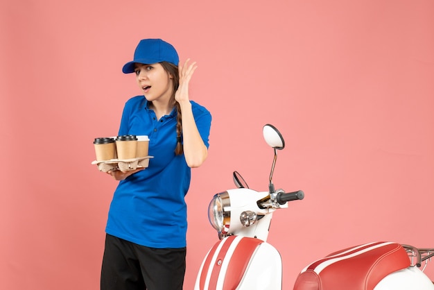 Vorderansicht des Kuriermädchens, das neben dem Motorrad steht und Kaffee und kleine Kuchen hält und den letzten Klatsch auf pastellfarbenem Pfirsichhintergrund hört