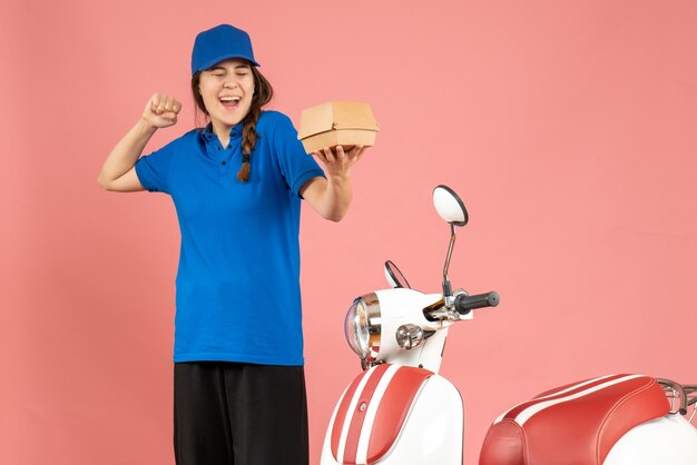 Vorderansicht des lächelnden Kuriermädchens, das neben dem Motorrad steht und Kuchen auf pastellfarbenem Pfirsichhintergrund hält