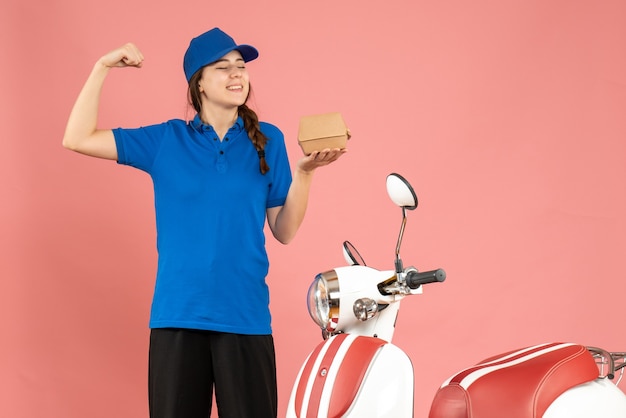 Vorderansicht des lächelnden Kuriermädchens, das neben dem Motorrad steht und Kuchen hält, der muskulös auf pastellfarbenem Pfirsichhintergrund zeigt