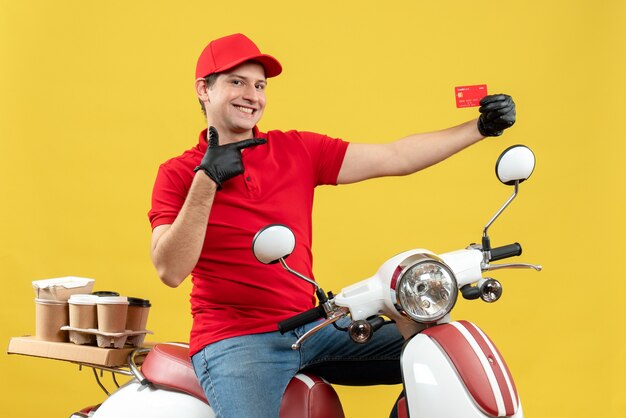Vorderansicht des lächelnden Kuriermannes, der rote Bluse und Huthandschuhe in der medizinischen Maske trägt, die Bestellung sitzend sitzt auf Roller zeigend Bankkarte
