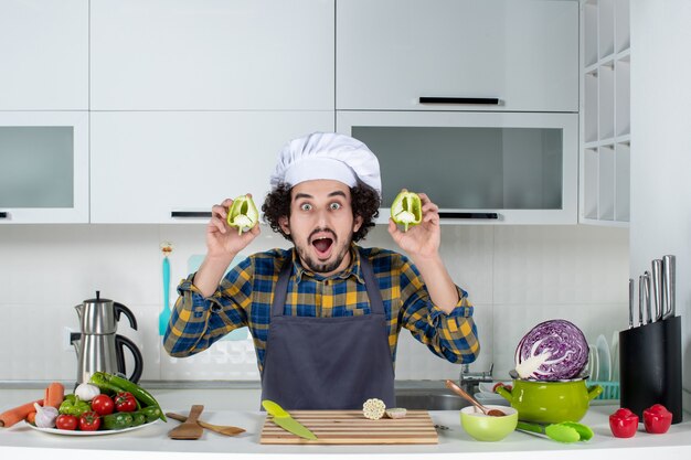 Vorderansicht des lächelnden männlichen Kochs mit frischem Gemüse und Kochen mit Küchengeräten und mit geschnittenen grünen Paprikaschoten in der weißen Küche