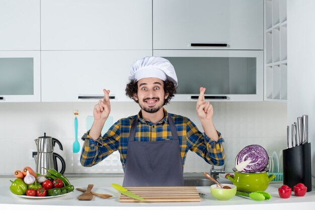 Vorderansicht des lächelnden männlichen Kochs mit frischem Gemüse und Kochen mit Küchengeräten und überkreuzen der Finger in der weißen Küche