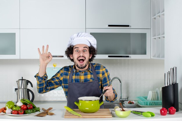 Vorderansicht des lächelnden und glücklichen männlichen Kochs mit frischem Gemüse, das Fertiggerichte schmeckt und in der weißen Küche eine Brillengeste macht