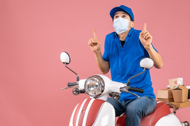 Vorderansicht des Lieferers in medizinischer Maske mit Hut, der auf dem Roller sitzt und auf pastellfarbenem Pfirsichhintergrund zeigt