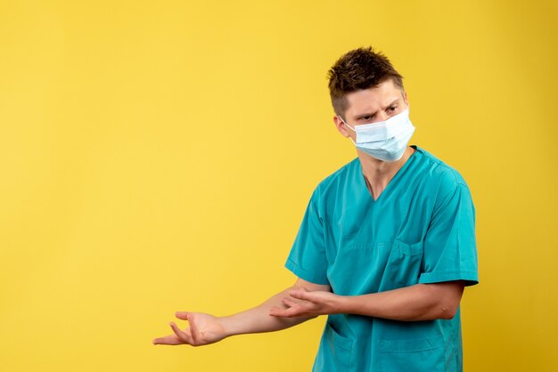 Vorderansicht des männlichen Arztes im medizinischen Anzug mit steriler Maske auf gelber Wand