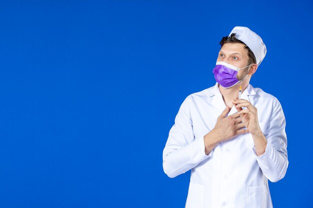 Vorderansicht des männlichen Arztes im medizinischen Anzug und in der lila Maske, die Injektion auf Blau hält