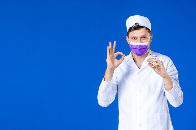 Vorderansicht des männlichen Arztes im medizinischen Anzug und in der lila Maske, die Injektion auf Blau hält