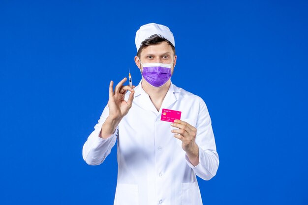 Vorderansicht des männlichen Arztes im medizinischen Anzug und in der lila Maske, die Injektion und Kreditkarte auf Blau hält