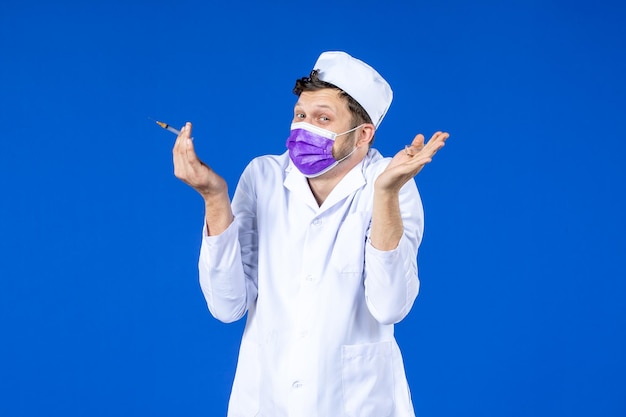 Vorderansicht des männlichen Arztes im medizinischen Anzug und in der Maske, die Impfstoff und Injektion auf Blau halten