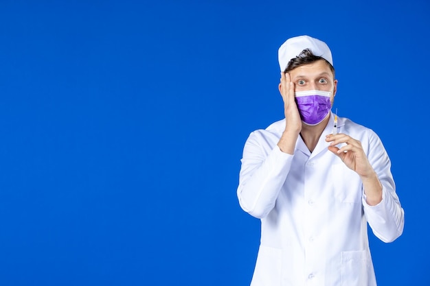 Vorderansicht des männlichen Arztes im medizinischen Anzug und in der Maske, die Injektion auf Blau hält