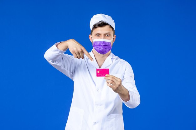 Vorderansicht des männlichen Arztes im medizinischen Anzug und in der Maske, die Kreditkarte auf Blau hält