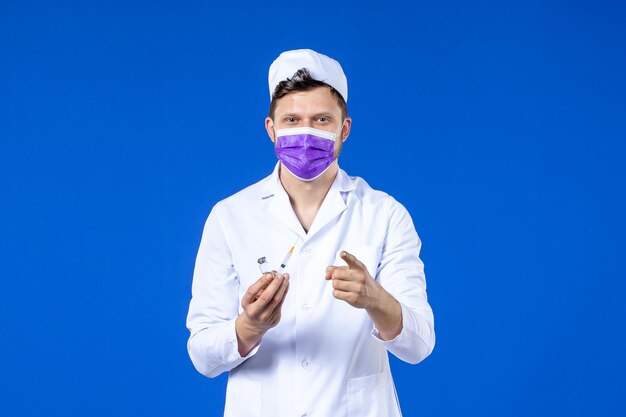 Vorderansicht des männlichen Arztes im medizinischen Anzug und in der Maske mit Injektion und Impfstoff auf Blau