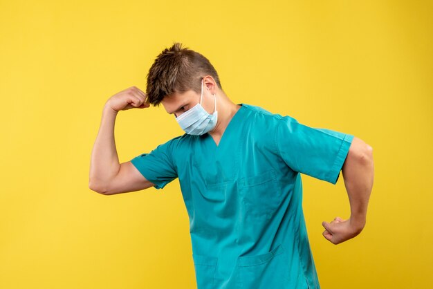 Vorderansicht des männlichen Arztes im medizinischen Anzug und in der sterilen Maske auf gelber Wand