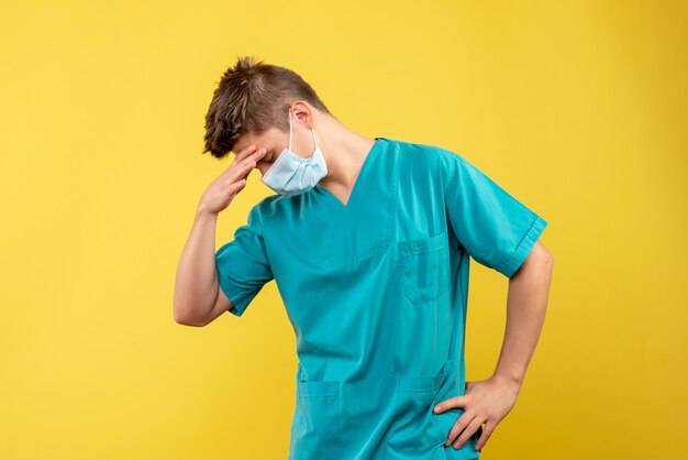 Vorderansicht des männlichen Arztes im medizinischen Anzug und in der sterilen Maske auf gelber Wand