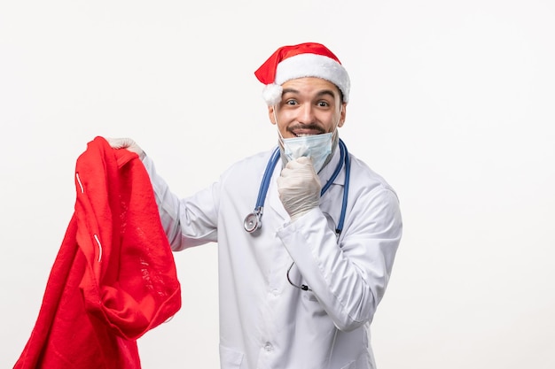 Vorderansicht des männlichen Arztes in Maske mit Geschenktüte an weißer Wand