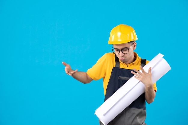 Vorderansicht des männlichen Baumeisters in der gelben Uniform und im Helm mit Plan auf blauer Wand