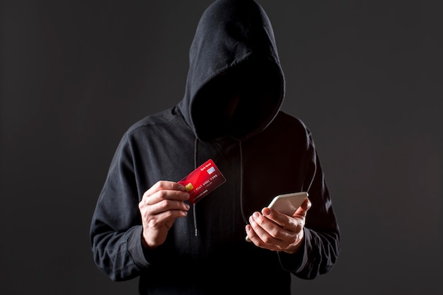 Vorderansicht des männlichen Hackers, der Smartphone und Kreditkarte hält