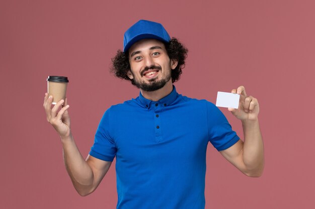 Vorderansicht des männlichen Kuriers in blauer Uniform und Kappe mit Kaffeetasse und Karte der Lieferung auf seinen Händen an der rosa Wand