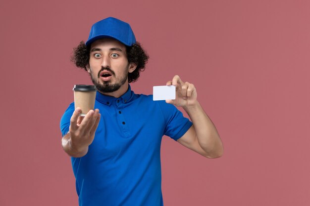 Vorderansicht des männlichen Kuriers in blauer Uniform und Kappe mit Kaffeetasse und Karte der Lieferung auf seinen Händen an der rosa Wand
